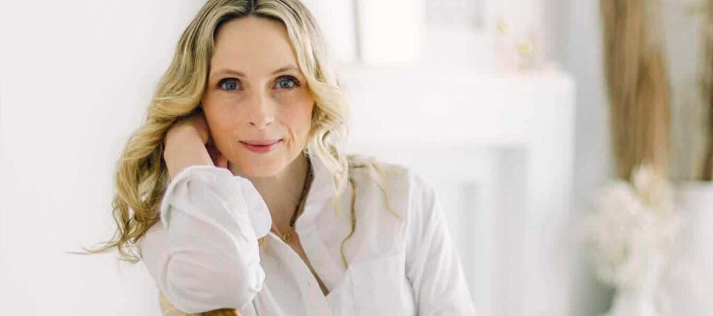 Stephenie Farrell, a blonde woman wearing a white blouse and blue jeans, leans sideways on a chair with her arm supporting her head. Stephenie Farrell is an integrative trauma recovery specialist based in Hamilton, Ontario, Canada.